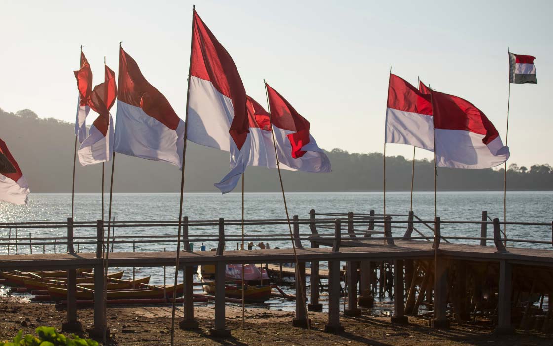 Indonesische Flaggen am Ufer eines Sees. Im Hintergrund liegt ein Steg, mehrere kleine Boote und am Horizont erstreckt sich eine Landzunge